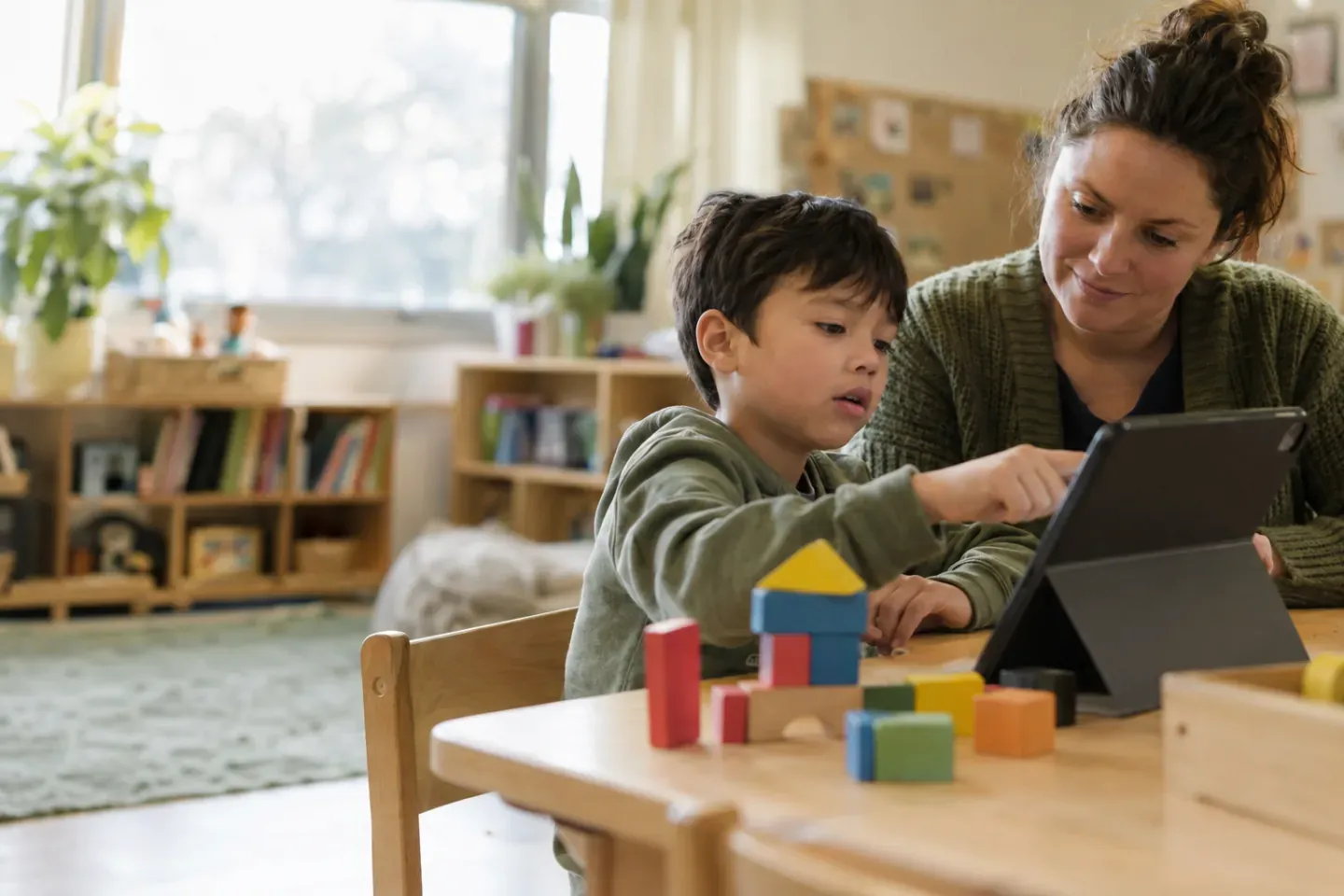 A student uses a tablet beside an adult at a classroom table with colorful building blocks.